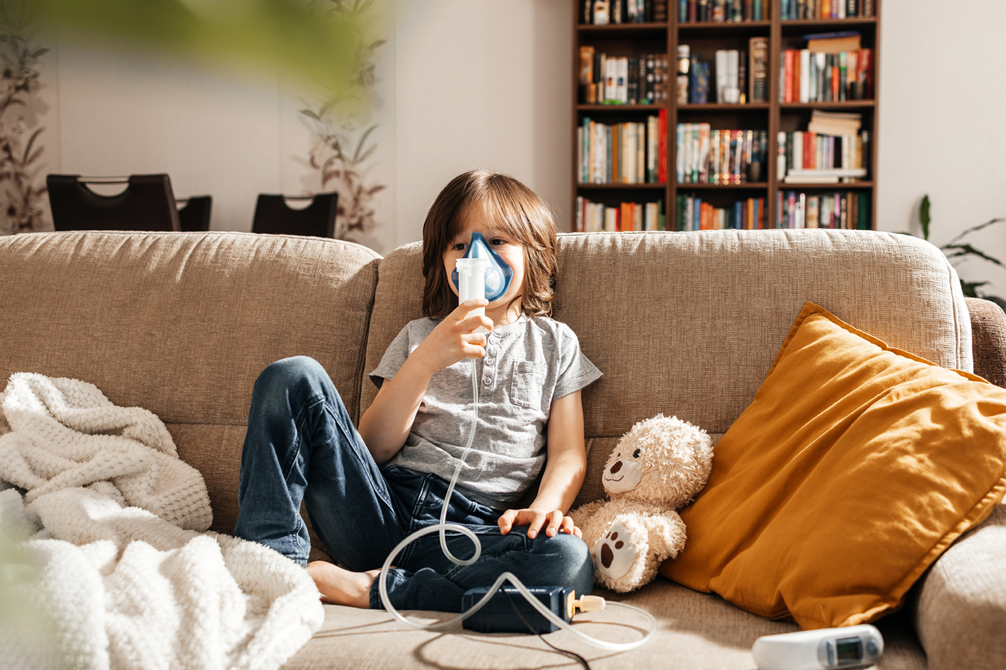 A little boy makes an inhalation at home sitting on the couch using a nebulizer and a mask.